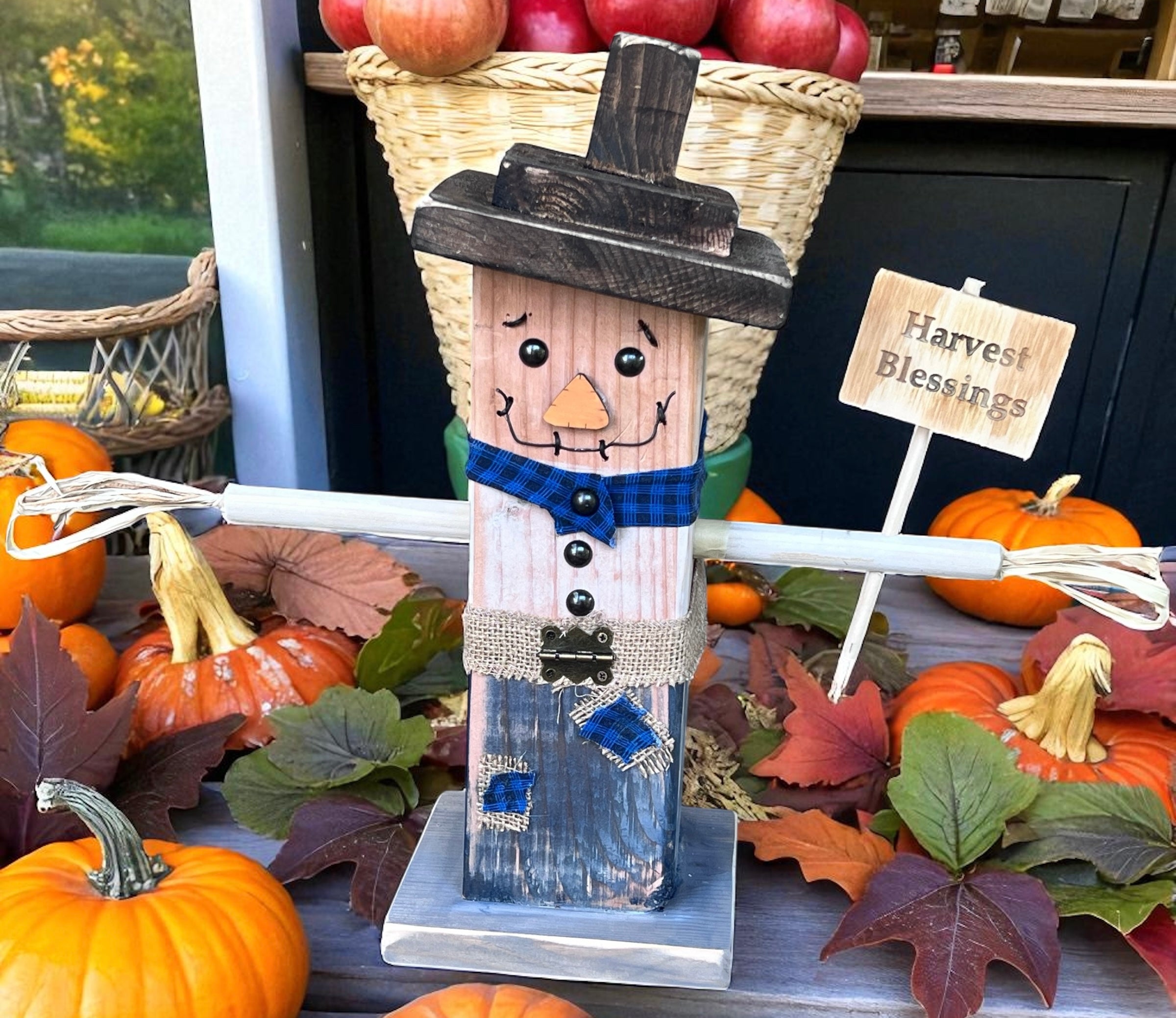 Handmade wooden scarecrow with a painted rustic finish and scarf, displayed on a farmhouse porch with fall pumpkins and crates.