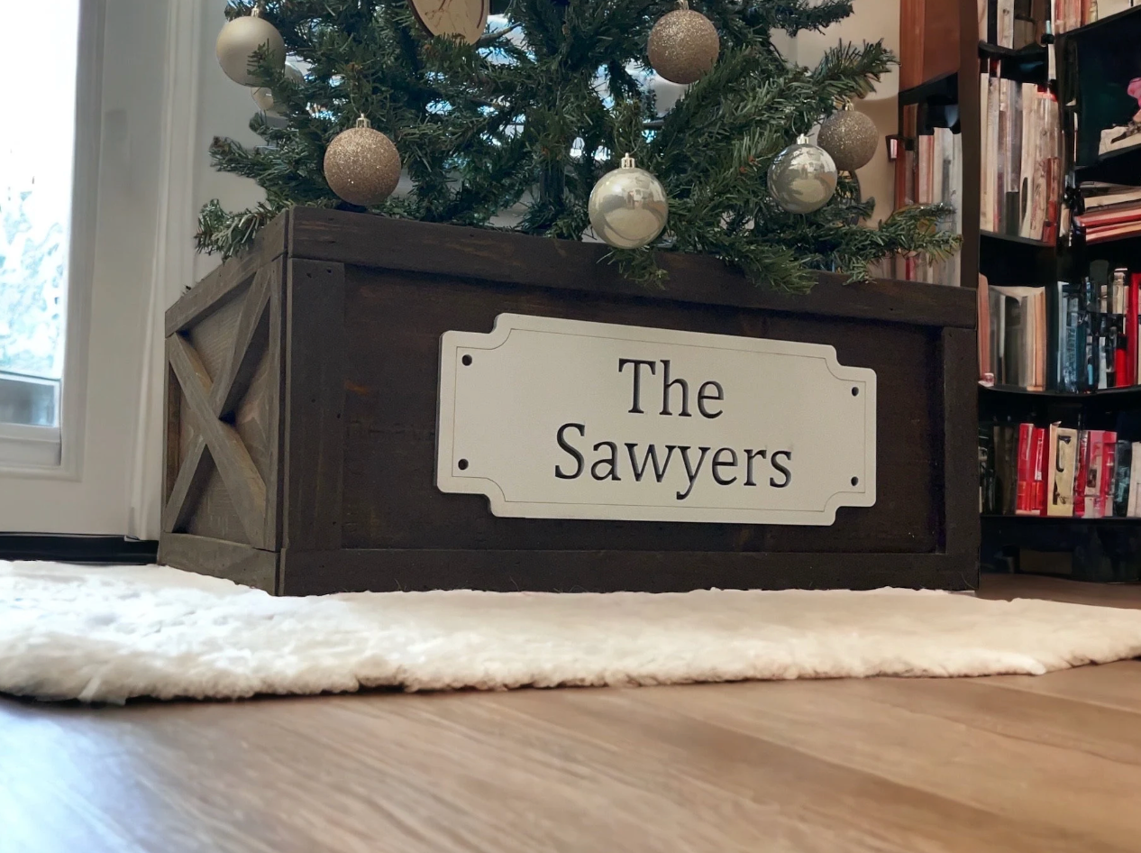 house-style wooden Christmas tree collar with custom name sign, stained brown, displayed under a Christmas tree.