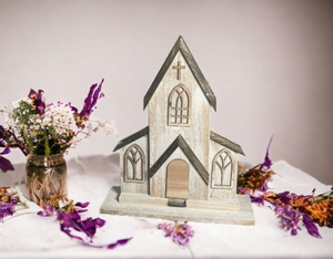 Rustic cedar church decor shown in a farmhouse-style display with purple and white flowers. The two-piece handcrafted design features arched windows, a cross, and natural wood roof accents. Perfect for weddings, religious gifts, or home faith decor.