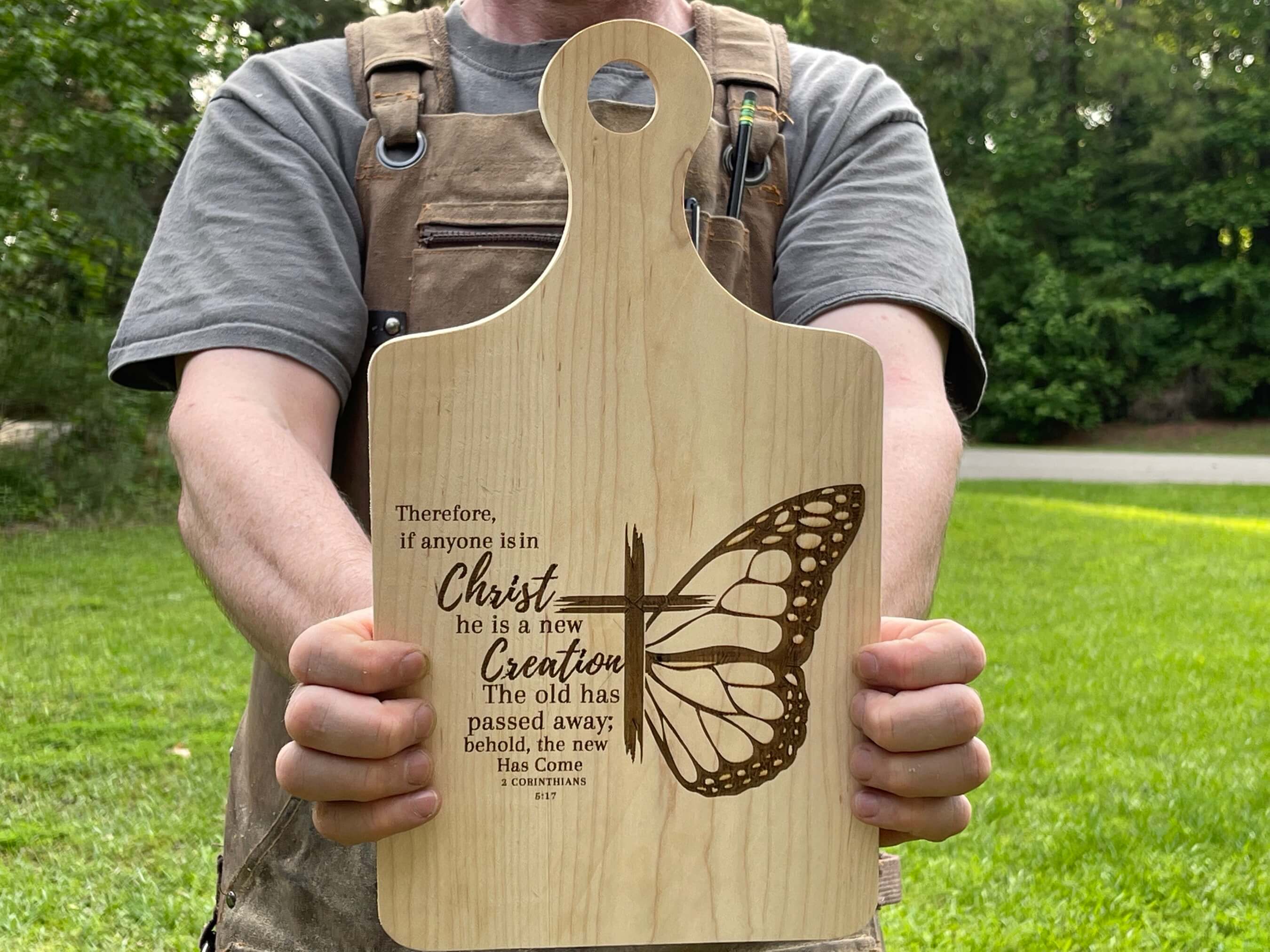 Closer view of the Christian cheese board. The cutting board is shown being held by someone outside. On the board is the bible verse 2 Corinthians 5:17. See more at SawyerCustomCrafts.com