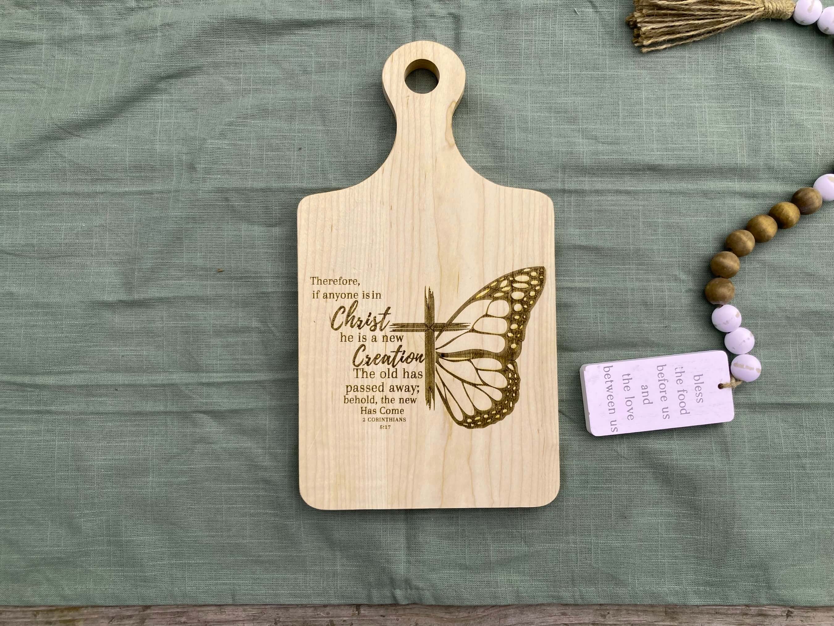 Jesus Cutting board Gift Idea. Here the cutting board is shown on a green background with some decoration on it. The cutting board says 2Cor. 5:17 Therefore, if anyone is iin Christ, he is ja new creation.2 kThe old has passed away; behold, the new has come.. sawyercustomcrafts.com
